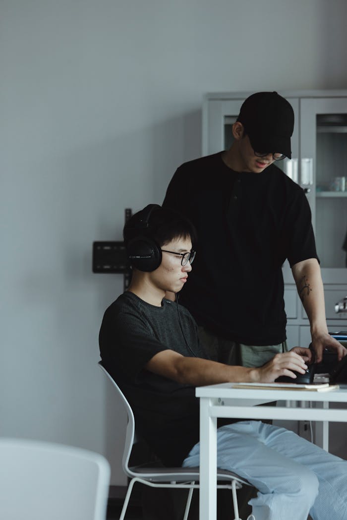 Two young men working together at a desk with a laptop and headphones in a modern office setting.